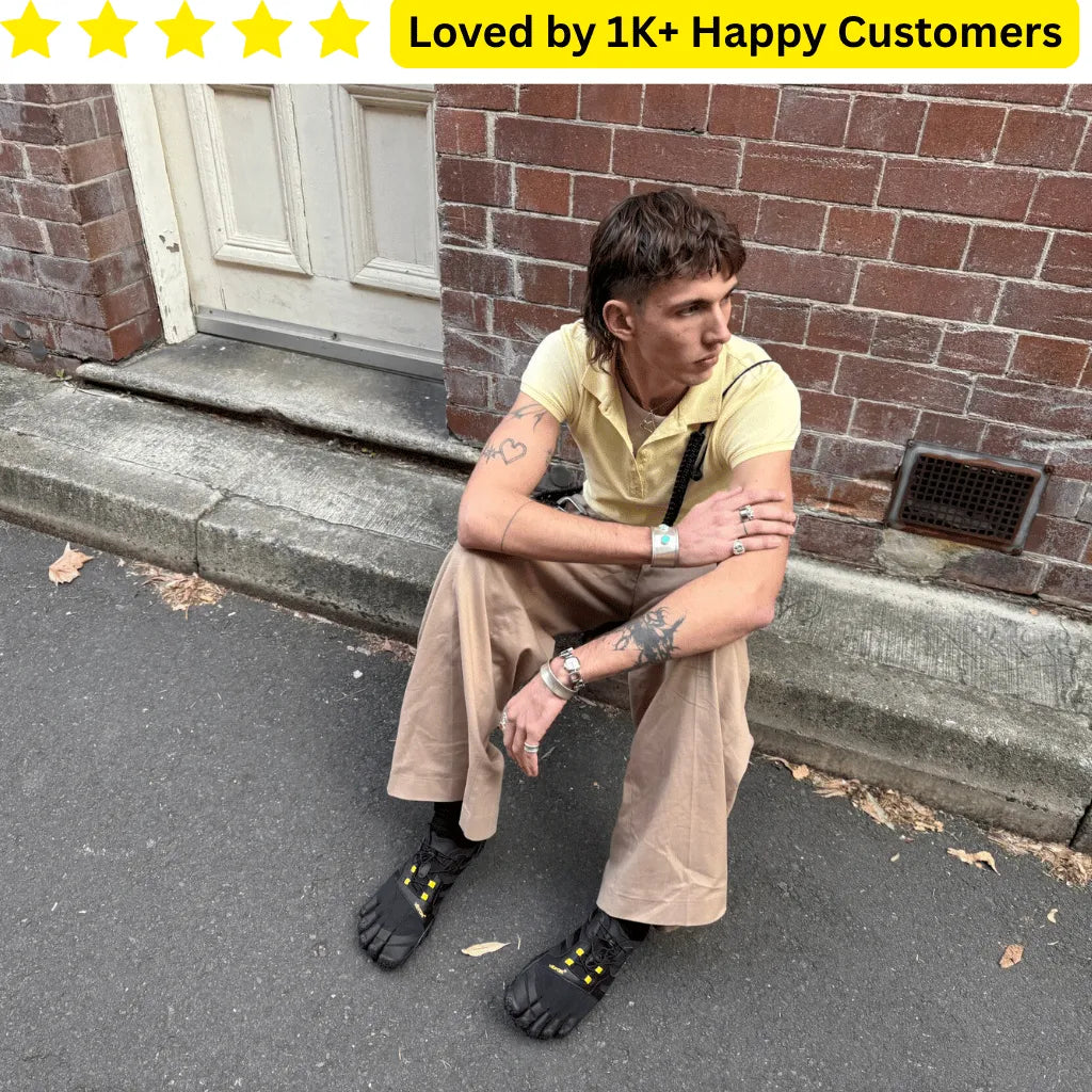 Person sitting on a step wearing tan pants and black shoes with yellow accents, with a brick wall and door in the background. Goth Tattoo – Upload Your Artwork & Get in 3 Days