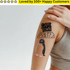 Tattoo of a penguin and 'Cape Town' on a person's arm with a hand holding it, on a light gray background.