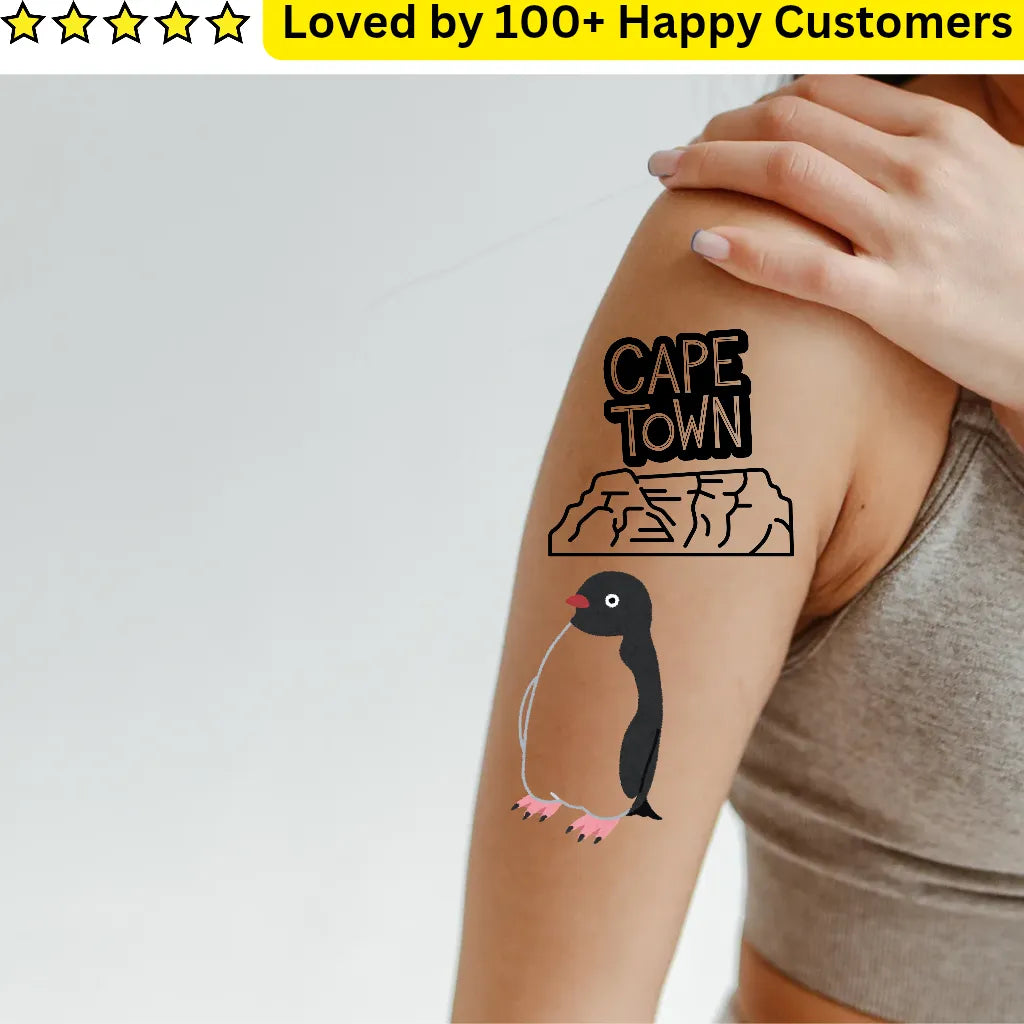 Tattoo of a penguin and 'Cape Town' on a person's arm with a hand holding it, on a light gray background.
